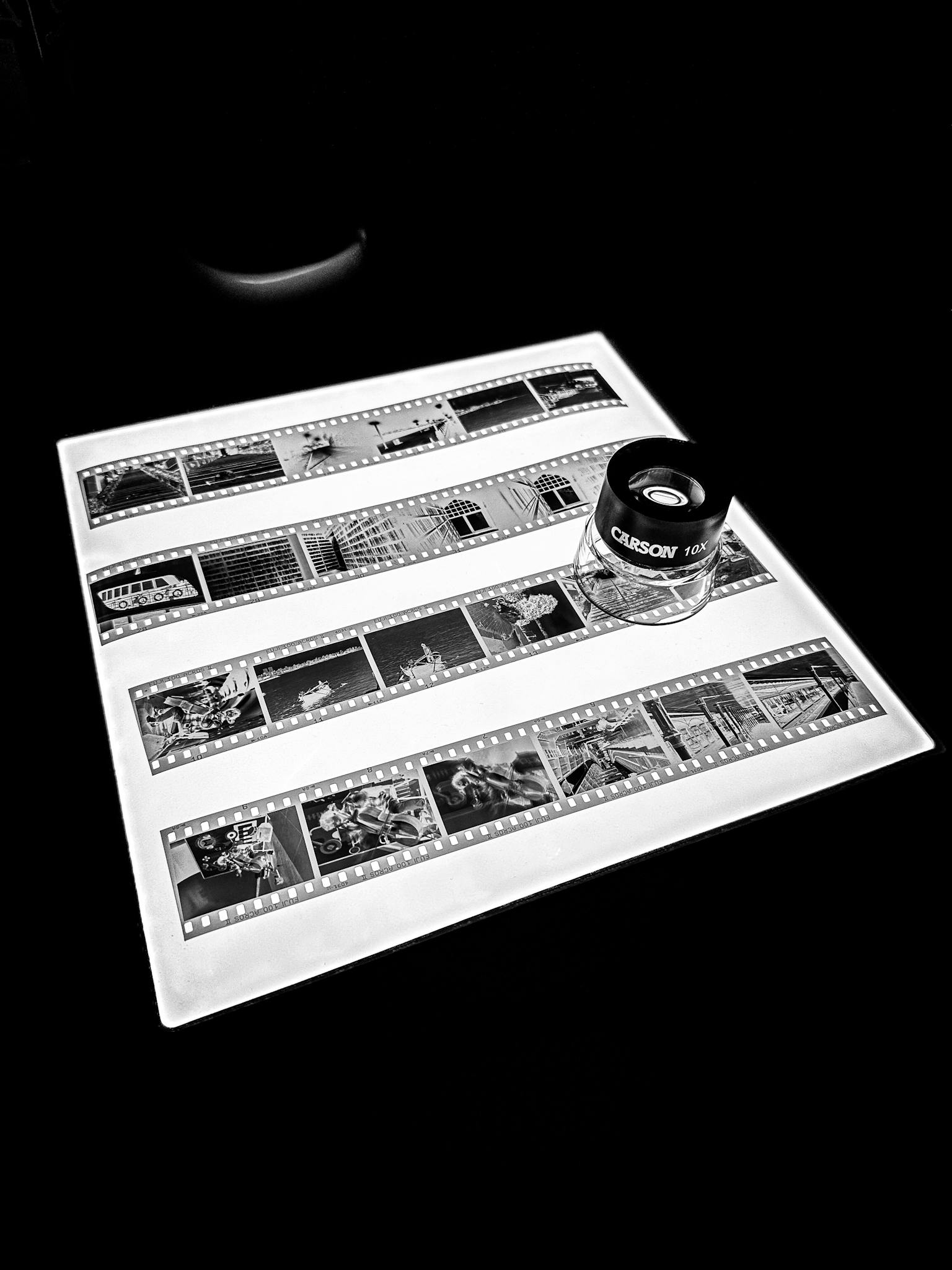 Schwarzweiss-Filmnegative auf einem Leuchttisch, betrachtet mit einer Präzisions-Lupenlupe für die sorgfältige Analyse analoger Fotografie.